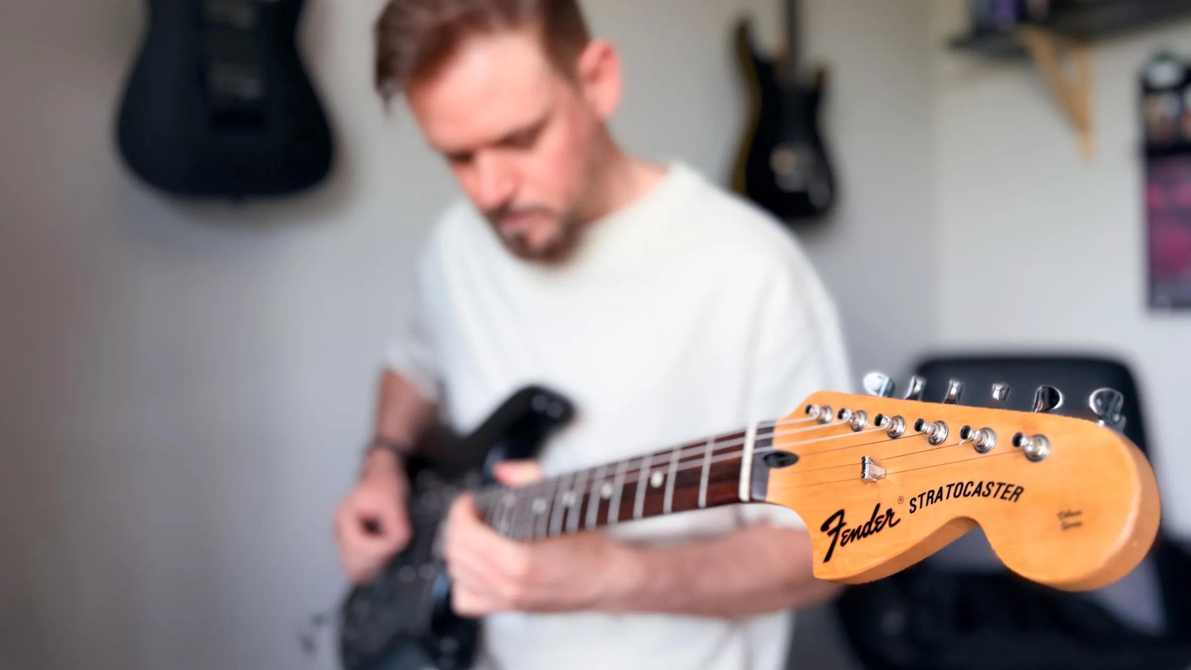A Fender Stratocaster headstock in sharp focus while a guitarist plays a black electric guitar, with more guitars hanging on the wall behind.